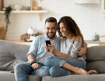 A smiling couple looks at a smartphone while sitting on a gray couch